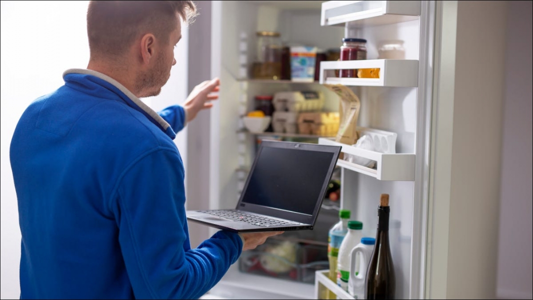 Человек, держащий ноутбук перед открытым холодильником.